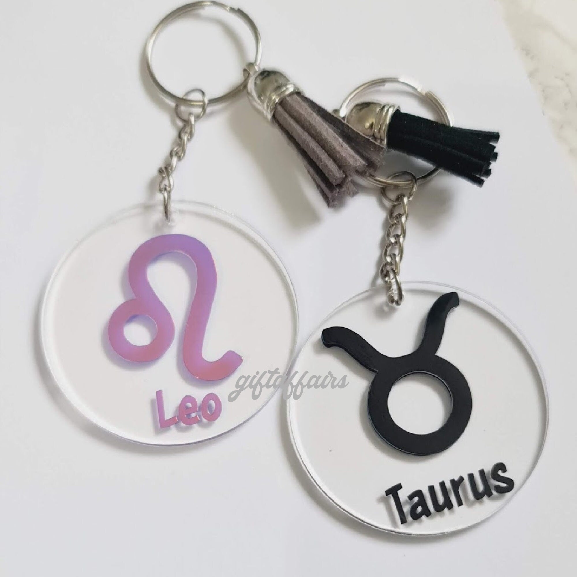 Two keychains with zodiac symbols, one pink 'Leo' and one black 'Taurus', on a white background.